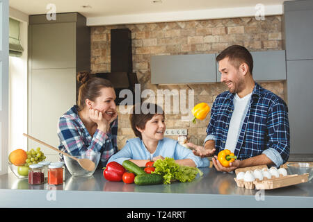 Mère Père et fils à la maison debout dans la cuisine ensemble woman and boy leaning on table looking at man juggling paprikas smiling curieux Banque D'Images