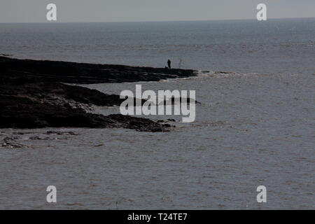 Un pêcheur solitaire se distingue avec sa canne et à la ligne à la fin d'un éperon promontary avec le clapotis des vagues à ses pieds sur une journée ensoleillée mais cool. Banque D'Images