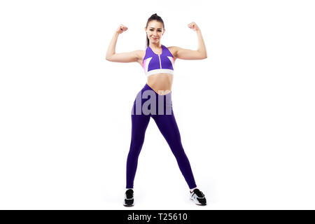 Une femme brune coach sportif dans une salle de sport et haut court violet leggings montre biceps et sourires sur un fond blanc en studio isolé Banque D'Images