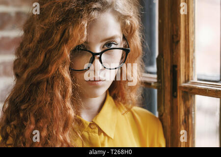 Portrait of smiling curly ginger girl à la chemise jaune portant des lunettes, debout près de la fenêtre au loft et placement à sérieusement pour l'appareil photo. Banque D'Images