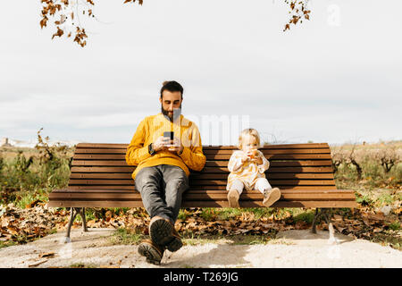 Père et fille assise sur un banc dans le parc en automne, le père à l'aide de smartphone, fille de manger une pomme Banque D'Images