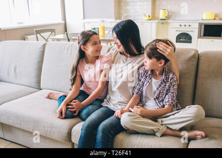 La mère est assise sur un canapé avec ses enfants. Elle est les étreignant. Femme regarde sa fille. Elle détient la main sur la tête du garçon. Heureux les enfants sont à la recherche de maman Banque D'Images