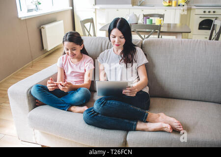 Les filles sont assis sur un canapé et lecture. Femme fille tout en tablette dispose d'un téléphone dans les mains. Ils ont l'air concentrés Banque D'Images