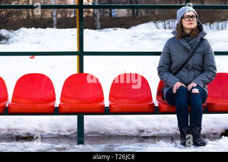 Une belle et jeune fille avec des lunettes est assis sur un banc de chaises rouges. Elle regarde pensivement le ciel avec les mains jointes sur ses genoux. Banque D'Images