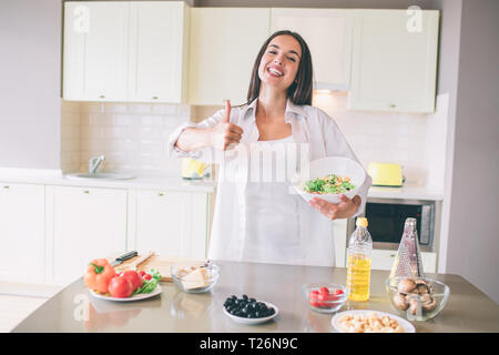 Heureuse jeune fille se tient et montre son gros pouce vers le haut. Elle a l'air sur la caméra et sourit. La fille aussi est titulaire d'bol avec de la salade. Il y a beaucoup d'ingrédients sur la table Banque D'Images