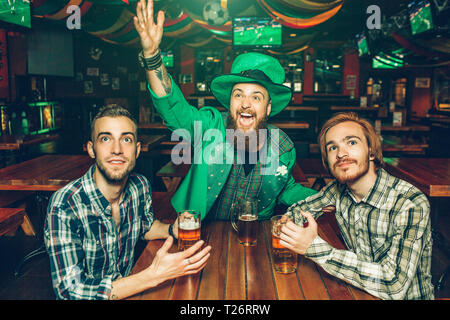 Heureux heureux jeune homme en costume vert forme avec les mains et parvenir à l'avant. Deux autres hommes s'asseoir à tabe et tenir la bière de tasses Banque D'Images