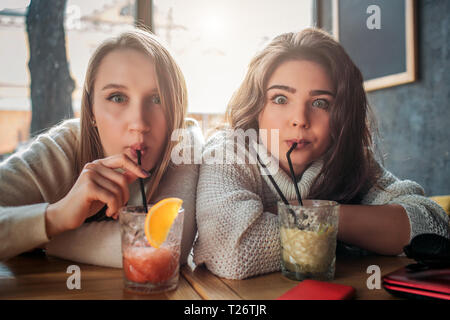 Les jeunes femmes s'assied à table et de boire des cocktails à travers la paille. Ils regarde droit. Modèles semblent drôles. Ils s'asseoir à table Banque D'Images