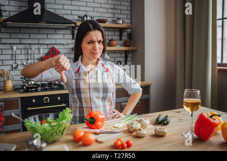 Jeune femme malheureuse s'asseoir à table dans la cuisine. Elle regardez sur appareil photo et maintenez le pouce vers le bas gros. Femme en colère. Légumes sur table. Le verre de vin sur 24 Banque D'Images