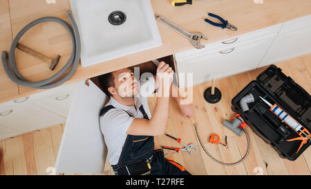 Plombier jeune regarder sous évier dans la cuisine. Il a mis le nouveau flexible. Outils avec boîte ouverte sur l'étage. Clé à tuyau flexible et des tenailles sur 24 Banque D'Images