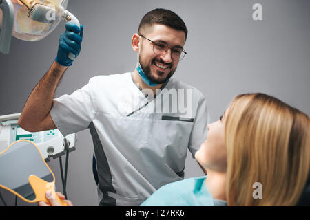 Photo de heureux dentiste s'asseoir à côté client et regarder la regarder. Il sourire et tenir la lampe. Femme assis dans fauteuil et regarder un dentiste. Banque D'Images