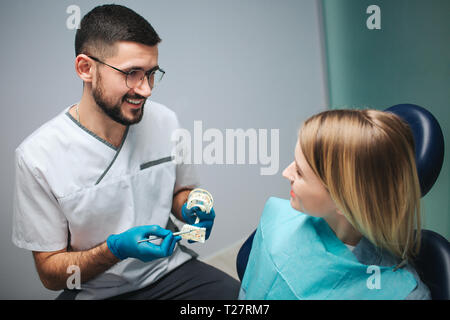 Jeune dentiste nice joyeuse attente mâchoire artificielle avec dents et point sur eux en dentisterie. Il regarde female client et sourire. Positive young woman Banque D'Images