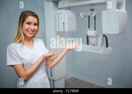 Positive heureux jeune dentiste en salle de radiographie et point sur la machine. Seul en robe blanche. Banque D'Images