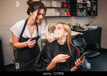 Coiffeur professionnel avec des ciseaux et belle clientèle féminine coupe de décider ce qui à faire pendant la recherche de téléphone. Coupe de l'évolution de la beauté Banque D'Images