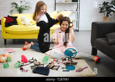 Heureux deux adolescentes s'amuser dans la chambre. Ils sont assis et debout sur le tapis. Fille blonde les cheveux de son amie de curling. Brunette faire un miroir et regardez dans mir Banque D'Images