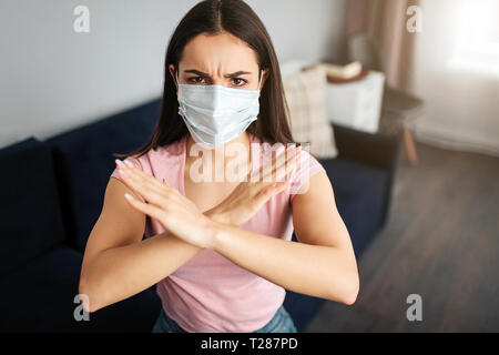 Jeune femme sérieuse s'asseoir sur un canapé. Elle a couvert la bouche avec un masque. Modèles est malade. Elle montre un panneau d'arrêt avec les mains. Jeune femme en colère Banque D'Images