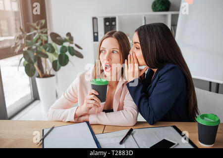 Deux jeunes femmes s'asseoir ensemble dans la salle à table. Brunette murmure à l'oreille d'une autre personne de sexe féminin. Ils potins. Fille blonde tenir tasse de café Banque D'Images