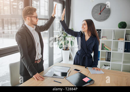Le travail d'équipe de jeune homme et femme. Ils se tiennent dans la chambre et donner une haute-cinq de l'autre. Les gens sourire. Ils ont l'air heureux et satisfaits Banque D'Images