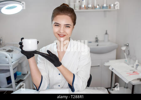 Beau jeune coiffeur tenir pot de crème pour le visage. Il voit sur l'appareil-photo sourire et poser. S'asseoir dans le modèle prix. Pot de crème point sur elle. Banque D'Images