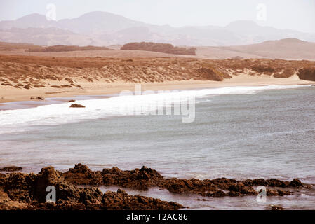 Rivage rocheux surplombant l'océan avec des vagues se brisant sur la plage de sable fin et au-delà de montagnes brumeuses sous un ciel brumeux lumineux. Banque D'Images