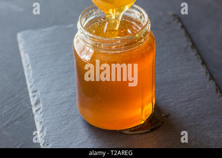 Le miel en pot de verre isolé sur fond d'ardoise noire - pouring Banque D'Images