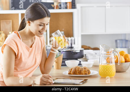 Smiling Pretty woman looking at mobile phone et holding verre de jus d'orange pendant le petit-déjeuner dans une cuisine. Banque D'Images