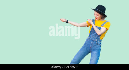 Vue côté profil portrait de jeune fille en bleu denim hipster salopette, chemise jaune et noir hat standing avec fist ou tirer geste. l'indo Banque D'Images