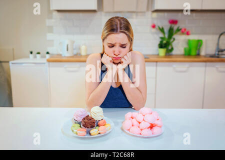 Lutte de régime. Jeune femme triste en bleu T-shirt choisit entre les légumes fruits frais ou des bonbons tout en le regardant dans la cuisine. Et en bonne santé Banque D'Images