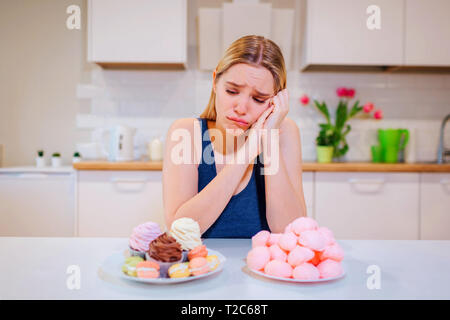 Lutte de régime. Jeune femme triste en bleu T-shirt choisit entre les légumes fruits frais ou des bonbons tout en le regardant dans la cuisine. Et en bonne santé Banque D'Images