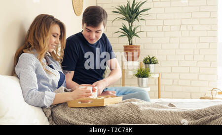 Belle fille blonde se trouve sur les oreillers guy sert le petit-déjeuner sur le bac au lit couple boit du café smiling Banque D'Images