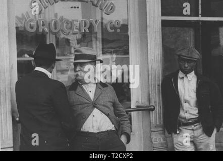 Photographie en noir et blanc de trois hommes debout devant l'mithland "Grocery Co ', y compris un homme mûr avec une grande moustache blanche et une chemise sale, qui s'entretient avec un homme portant un chapeau et costume, tandis qu'un troisième homme, portant un chapeau légèrement incliné et un noir, veste à glissière, s'appuie contre une colonne à l'extrême droite, regard vers la caméra, avec une expression sérieuse sur son visage ; situé à Smithland, Kentucky, USA ; photographié par Ben Shahn, sous le parrainage des membres de l'Administration de la sécurité à la ferme, en 1935. À partir de la Bibliothèque publique de New York. () Banque D'Images