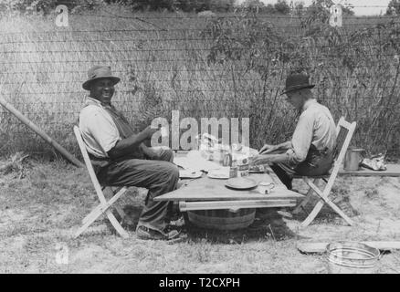 Photographie en noir et blanc de deux hommes, de profil, porter un chapeau et des vêtements de travail, assis dehors, sur des chaises en bois pliantes qui sont tirés jusqu'à une table en bois de fortune chargés de tasses, assiettes, et une variété de plats et de récipients de nourriture ; l'homme à gauche tient une tasse dans une main et se tourne vers la caméra avec un sourire bouche ouverte, l'homme à droite regarde vers le bas vers la nourriture qu'il prépare dans son assiette ; situé à Shelbyville, Kentucky, USA ; photographié par Ben Shahn, sous le parrainage des États-Unis' Farm Security Administration, août 1940. À partir de la Bibliothèque publique de New York Banque D'Images