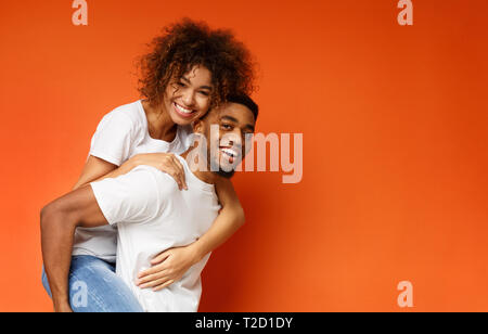 L'amour est tellement amusant ! Happy african-american man and woman riding piggyback, fond orange, de l'espace libre Banque D'Images