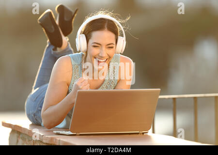 Heureux girl wearing headphones regarder media sur l'ordinateur portable posé sur un balcon mur Banque D'Images