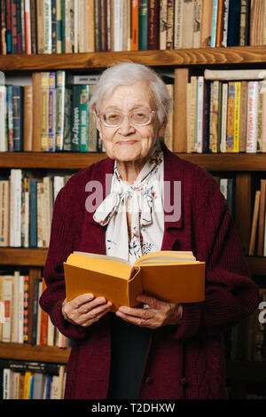 Portrait des personnes âgées gaies femme aux cheveux gris. La lecture, l'article avec réserve en mains près de bibliothèque à la maison Banque D'Images