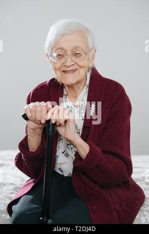 En particulier cheerful senior grey haired woman sitting on bed, la maintenant sur bâton de marche à la maison. Banque D'Images