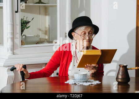 Bien respectable vêtus de noir mini hat grey haired woman fantaisie avec la canne est en train de lire son livre à table à dîner à la maison sur une journée de printemps ensoleillée. Banque D'Images