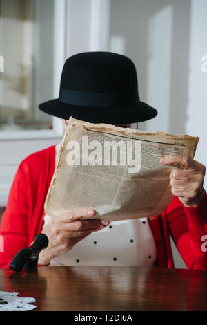 Vêtus de noir coloré mini hat grey haired woman fantaisie avec de la canne à sucre est la lecture d'un très vieux magazine papier miteux usé étroitement à face à dinn Banque D'Images