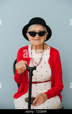 Bien respectable vêtus de noir mini chapeau et des lunettes de haute fantaisie femme aux cheveux gris avec de la canne à sucre est en appui sur une chaise à la maison sur une journée de printemps ensoleillée Banque D'Images