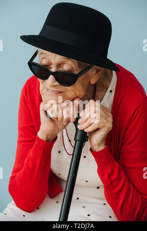 Bien respectable vêtus de noir mini chapeau et des lunettes de haute fantaisie femme aux cheveux gris avec bâton de marche est en appui sur une chaise à la maison sur une terrasse bien s Banque D'Images