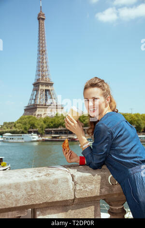 Femme élégante heureux dans l'ensemble des jeans avec tasse de café et de croissants, aller à des attractions contre vue claire de la Tour Eiffel et de la Seine Banque D'Images