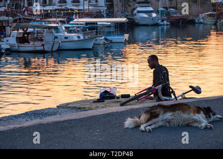 Kyrenia, Chypre - 27 novembre 2018 : Un homme africain se distingue par la mer sur le vieux port de Kyrenia à Chypre alors que son chien est en train de dormir sur l'aire Banque D'Images