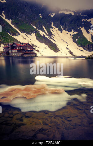 Un lac glaciaire avec un peu de glace capuchon fixé très haut au-dessus dans les montagnes avant la neige a fondu complètement avec une maison sur la rive en Roumanie Banque D'Images