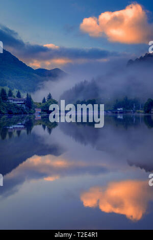 Matin brumeux sur un lac dans les montagnes avec quelques maisons sur la rive et nuages spectaculaires avec de beaux reflets dans l'eau Banque D'Images