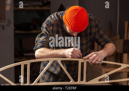 Le charpentier l'homme à chapeau et une chemise et marque les mesures avec les crayons points souhaités sur la planche de bois dans l'atelier, vue latérale, dans le bac Banque D'Images