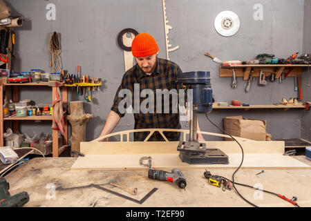 Un homme de chantier dans un chapeau et une chemise est une sculpture de bois sur une grande machine de forage en vue de côté, un atelier à l'arrière-plan beaucoup d'outils Banque D'Images