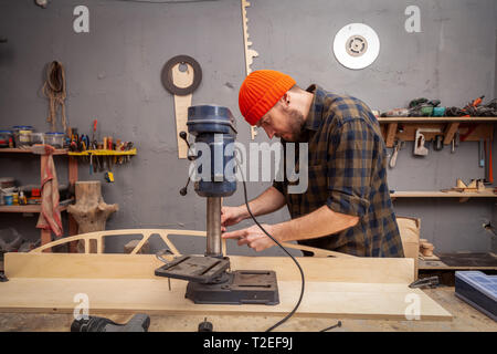 Un homme de chantier dans un chapeau et une chemise est une sculpture de bois sur une grande machine de forage en vue de côté, un atelier à l'arrière-plan beaucoup d'outils Banque D'Images