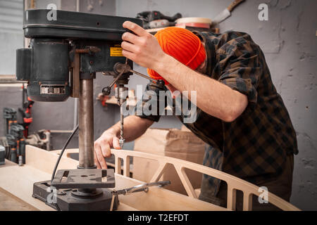 Un homme avec des vêtements de travail et d'une menuiserie hat est une sculpture de bois sur une grande machine de forage dans un atelier de lumière Banque D'Images