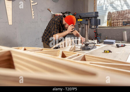 Un homme de chantier dans un chapeau et une chemise est une sculpture de bois sur une grande machine de forage en vue de côté, un atelier à l'arrière-plan beaucoup d'outils Banque D'Images