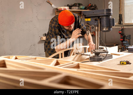 Un homme de chantier dans un chapeau et une chemise est une sculpture de bois sur une grande machine de forage en vue de côté, un atelier à l'arrière-plan beaucoup d'outils Banque D'Images