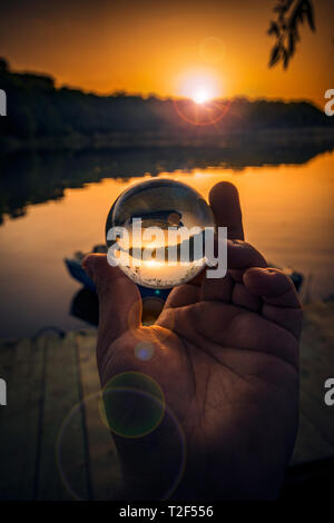 Shot conceptuel avec une main tenant une lensball utilisé pour les images inversées en raison d'un bateau de pêche sur un lac près d'un ponton au coucher du soleil Banque D'Images